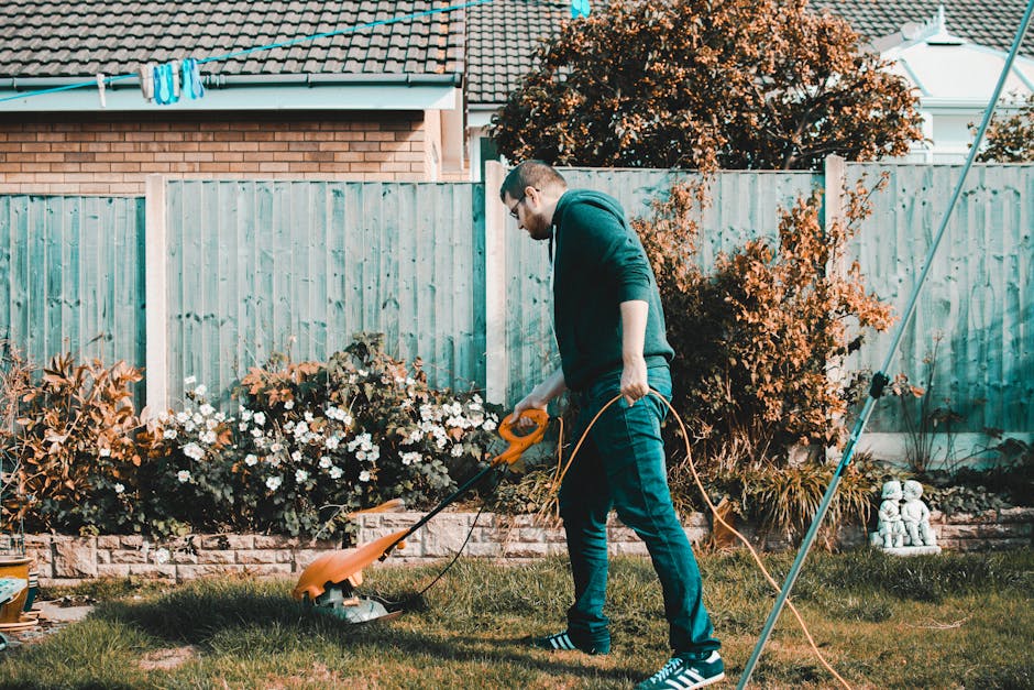 A man wearing glasses, a dark hoodie, and blue jeans is engaged in garden maintenance in a residential backyard. He is using an electric grass trimmer to cut the lush, green lawn, which has patches of